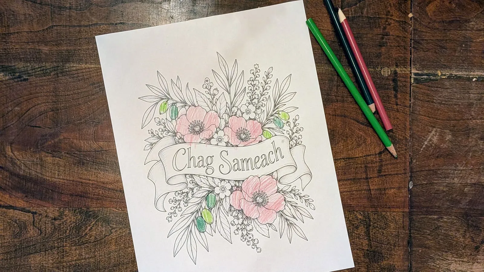 The finished Chag Sameach banner coloring page with soft pink blossoms and green leaves, colored pencils resting on a wooden table