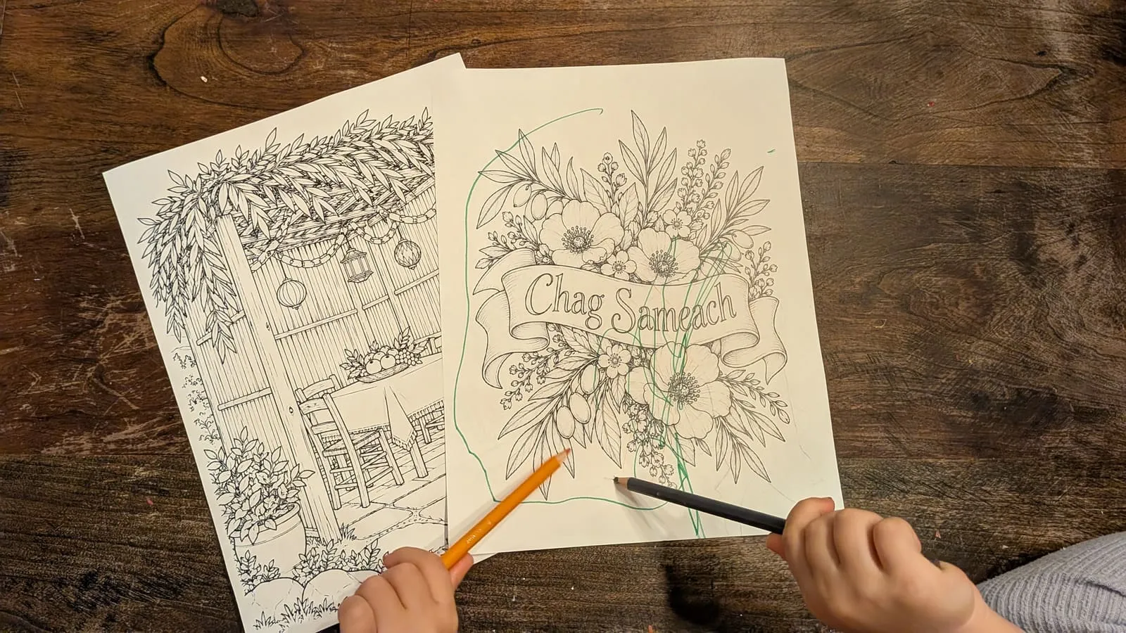 Two small hands with pencils coloring the Chag Sameach banner, the sukkah table coloring page tucked underneath