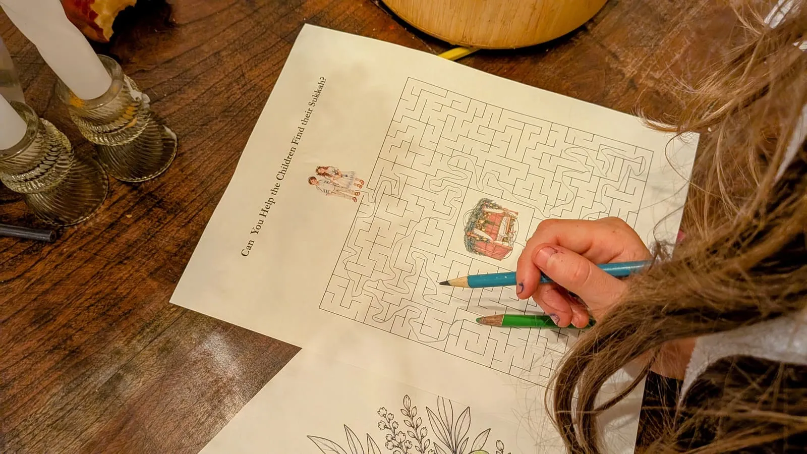 A child works the Help the Children Find Their Sukkah maze with a blue pencil, brass candlesticks glowing at the edge of the table