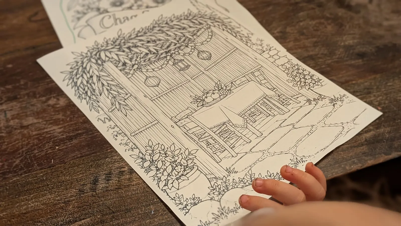 A toddler's hand resting on the sukkah table coloring page, branches and hanging fruit drawn along the roof