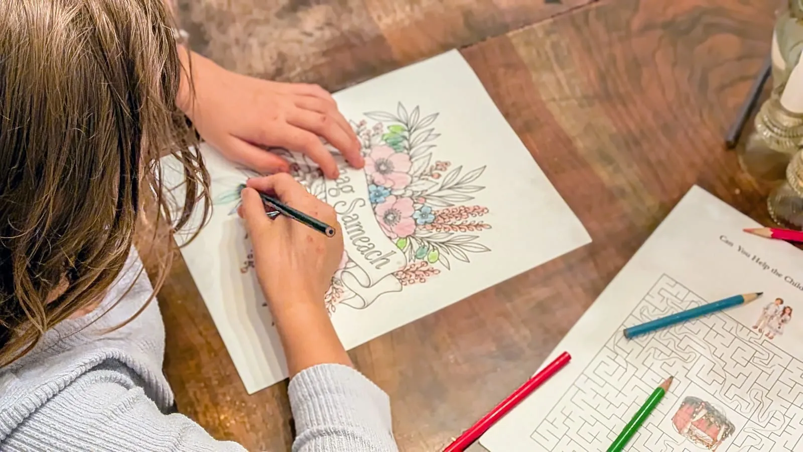 Seen from above, a child colors the Chag Sameach banner page in pinks and greens while a Sukkot maze waits beside it