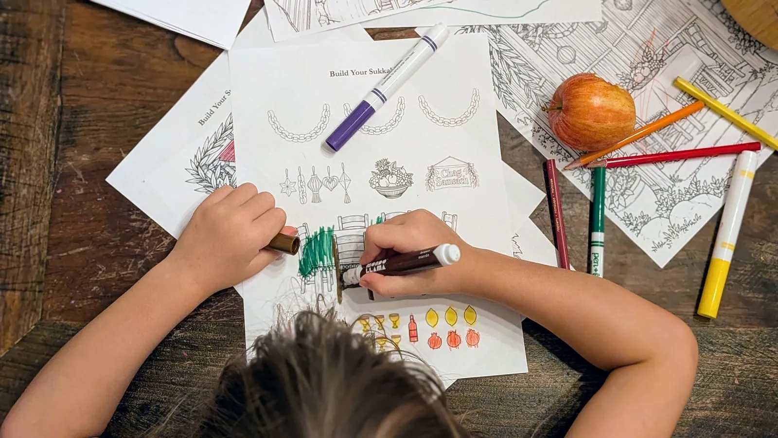 A child coloring the Build Your Sukkah decoration sheet with markers at a wooden table, an apple and a spread of colored pencils beside the page